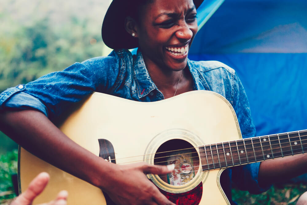 Photograph of a woman sat outdoors, smiling and strumming on an acoustic guitar.