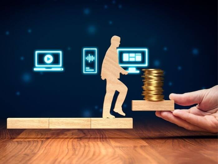 Wooden person stepping across ascending blocks, there are coins on the final step representing financial growth. In the background there are icons of a laptop, phone and PC, representing distribution.