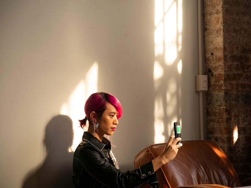 Photograph of a young person with bright pink hair, sat in a shadowy room they're holding up their phone and appear to be taking a photograph with it.