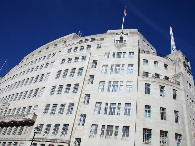 Photograph of the BBC Portland Place building, a large grey building with art decco features. The sky above is a bright blue.
