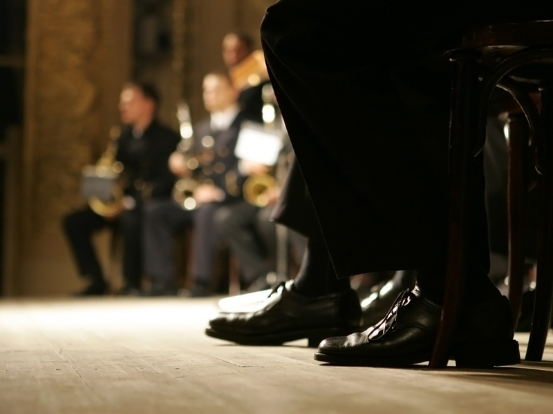 Photograph of a group of musicians sitting to play. The photograph is taken from the floor, and the primary focus is on the shoes of the musicians closest to the camera.