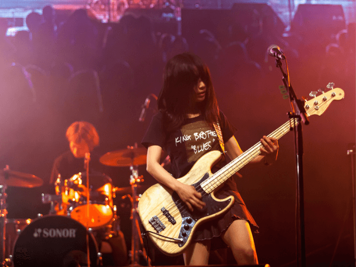 A female electric guitarist performing live on stage at a festival, with a drummer in the background and audience members onlooking.