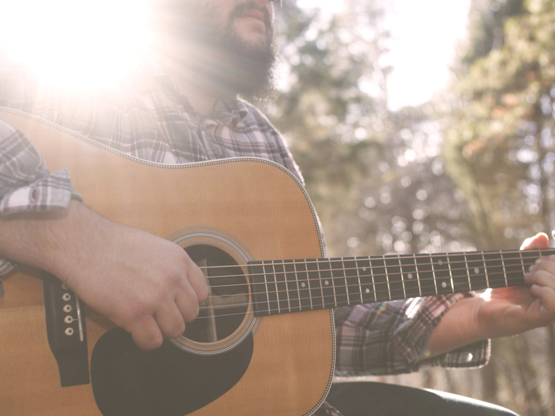 Photograph of a man playing an acoustic guitar, he is outdoors in the sunshine, and a sun flare covers his face.