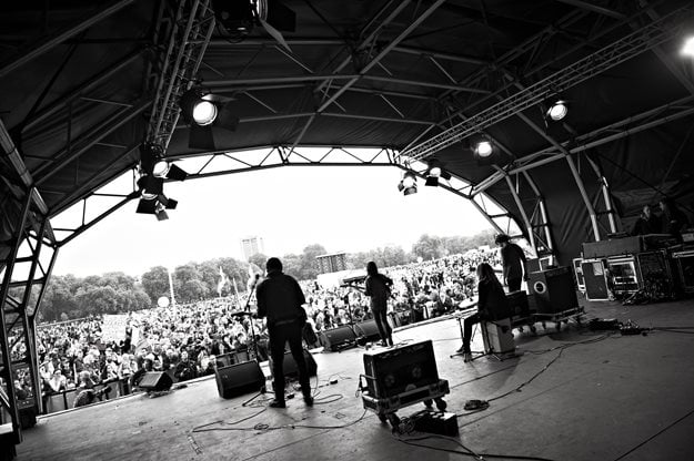 Black and white photograph from backstage at a festival. We can see a number of musicians playing on stage from behind, and a large outdoor audience facing inwards.