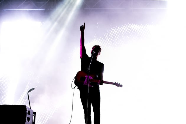 Musician silhouetted against a pale purple background, holding a guitar, one arm raised.