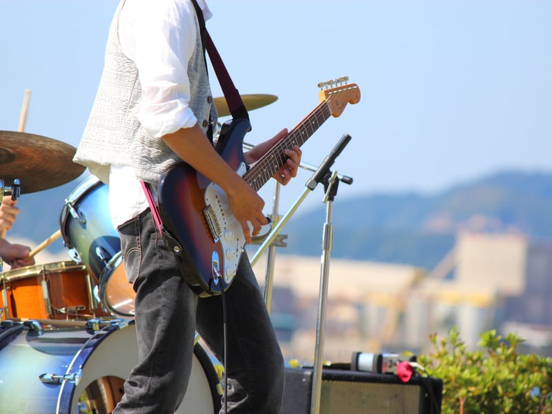 Photograph of a music event outdoors. The guitarist is gesturing dramatically from the stage with pick and electric guitar, and you can see a drum kit with a drummers hands behind.