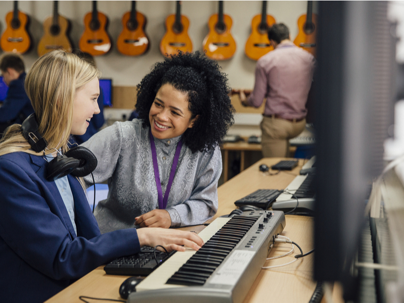 Photograph of a teacher working with a student in a classroom, the student is playing onto a keyboard and the teacher is smiling encouragingly.