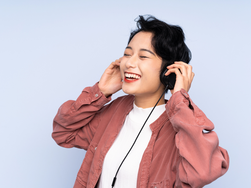 A young asian woman against a bright blue background is listening to music through headphones and smiling broadly